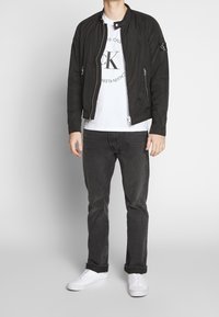 Svart zip-up bomberjacka med ribbad krage, med sidofickor och en logotypapplikation. Buren över en vit T-shirt och svarta jeans.