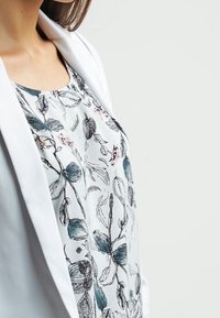 Blus med blommönster på vit bakgrund, med blå och rosa botaniska illustrationer, mjukt tyg, rund halsringning, lagd under en vit kavaj.