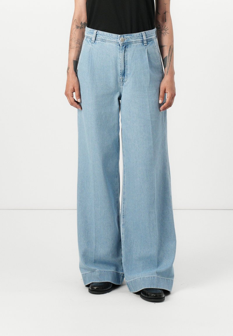 Pantalons en denim bleu clair à jambes larges avec une taille haute, dotés d'un bouton de fermeture et de lignes épurées. Le tissu a une texture lisse.