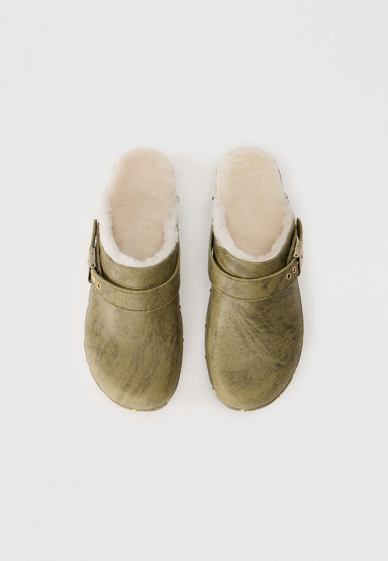 Grüne Lederclogs mit einem weichen, cremefarbenen Kunstfellfutter und einem dekorativen Riemen mit einer goldfarbenen Schnalle. Strukturiertes Oberflächendesign.