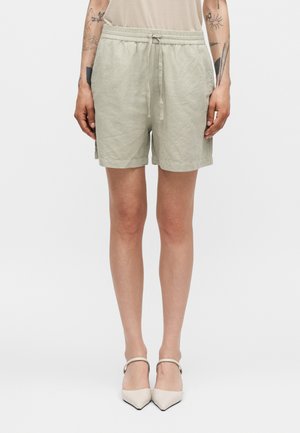 Personne portant un short beige clair en mélange de lin avec taille à cordon et poches latérales, associé à des talons blancs à bouts pointus avec brides à la cheville.