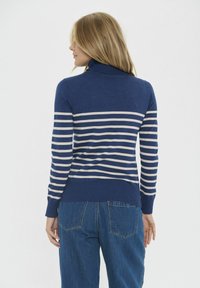 Pulover bleu marin cu guler înalt și dungi crem orizontale, cu manșete și tiv din tricot. Purtați-l cu blugi albaștri, văzut din spate.