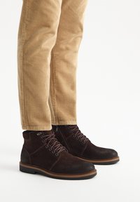 Brązowe zamszowe botki za kostkę z ciemnymi akcentami, sześciodziurkowym sznurowaniem, gumową podeszwą i kontrastującym jasnobrązowym przeszyciem. Połączone z beżowymi spodniami z kordurowej tkaniny.