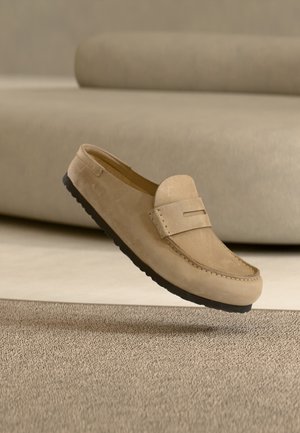 Mocassin en daim beige avec semelle noire posé sur un tapis beige texturé, avec un canapé beige assorti en arrière-plan.