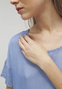 Vaaleansininen chambray-paita lyhyillä hihoilla, pyöreällä kaula-aukkolla, näkyvillä ompelu-yksityiskohdilla ja rentolla istuvuudella. Käsi lepää olkapäällä.