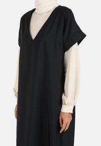 Black V-neck dress with short sleeves layered over a light beige long-sleeve top. Fabric appears smooth, with a relaxed fit and clean lines.
