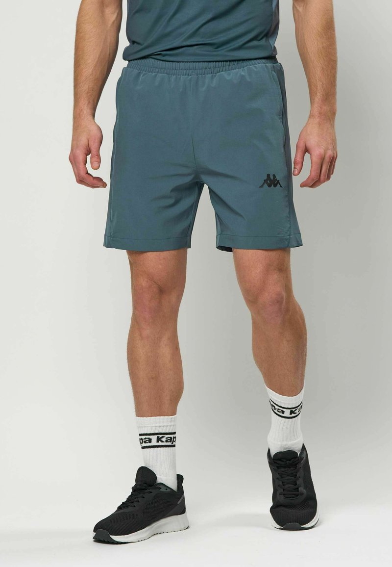 Shorts de sport pour hommes en bleu sarcelle, fabriqués en tissu léger. Taille élastique, poches latérales, et un accent de logo noir sur le côté inférieur gauche.