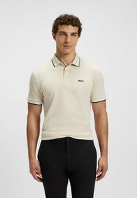 Polo beige con dettagli neri, maniche corte e tre bottoni. Presenta un logo sul lato sinistro del petto. Abbinato a pantaloni neri.