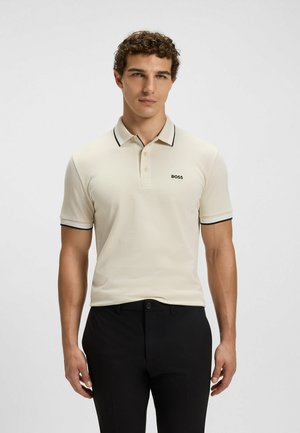 Polo beige con dettagli neri, maniche corte e tre bottoni. Presenta un logo sul lato sinistro del petto. Abbinato a pantaloni neri.