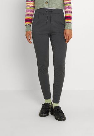 Kvinde iført mørkegrå bukser med snor, lysegrønne sokker, sorte loafers og en cropped grå cardigan med farverige mønstrede ærmer.