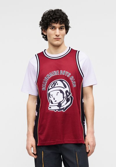 Νεαρός άνδρας με σγουρά μαλλιά, φοράει μια κόκκινη αμάνικη φανέλα "Billionaire Boys Club" πάνω από ένα λευκό T-shirt, στέκεται μπροστά σε απλό φόντο.
