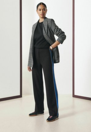 Vrouw draagt een grijs geruite blazer over een zwart topje, zwarte broek met wijde pijpen en blauwe zijstrepen, en zwarte sneakers, staand voor een witte achtergrond.