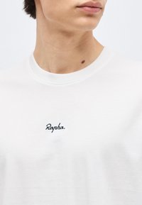 Gros plan d'une personne portant un t-shirt blanc à col rond avec le mot "Rapha." brodé sur la poitrine et un petit grain de beauté sur le cou.