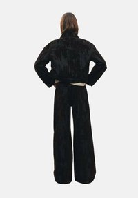 Completo di giacca in corduroy nero e pantaloni a gamba larga, con finitura testurizzata e design corto, visto da dietro su uno sfondo neutro.