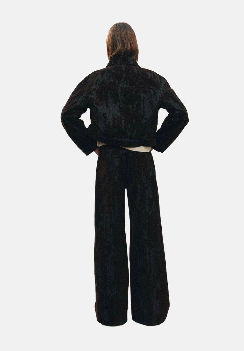 Completo di giacca in corduroy nero e pantaloni a gamba larga, con finitura testurizzata e design corto, visto da dietro su uno sfondo neutro.