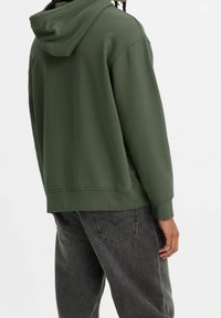 Personne portant un sweat à capuche vert foncé et un jean noir délavé, vue de dos sur un fond clair uni.