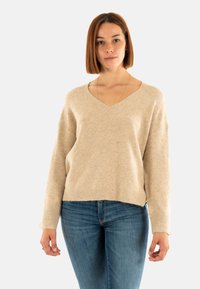 Mujer con cabello castaño liso hasta los hombros, vistiendo un suéter beige de cuello en V y jeans azules, de pie frente a un fondo claro y sencillo.