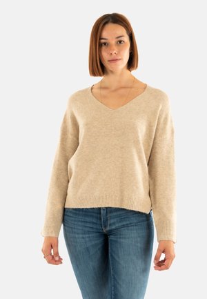 Femme aux cheveux bruns lisses de longueur épaule portant un pull beige en V et un jean bleu, se tenant devant un fond clair uni.