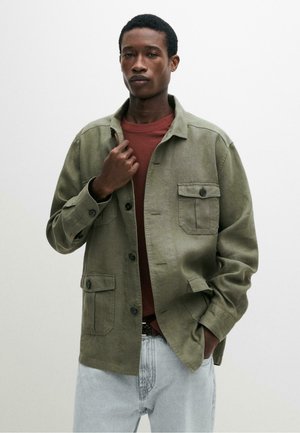 Homme portant une veste boutonnée vert olive sur une chemise rouge et un jean bleu clair, posant avec une main sur le col de la veste et l'autre dans la poche.