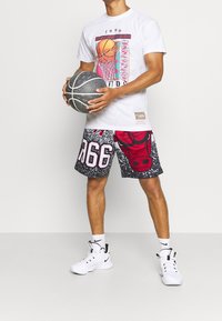 Mies, joka pitää mustaa koripalloa, päällään valkoinen T-paita, jossa on koripallokori -graafikka, sekä mustat ja punaiset Chicago Bulls -shortsit valkoisilla lenkkareilla.