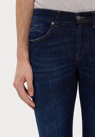 Jeans in denim blu scuro con un classico design a cinque tasche, dettagli in ottone e cuciture visibili. Texture liscia, forma aderente.