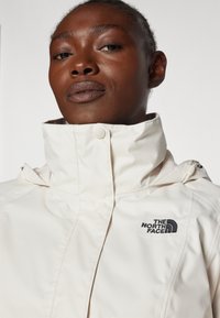 Giacca beige chiaro con colletto alto e logo nero "The North Face". Texture liscia e design elegante, con chiusura a bottoni e cappuccio.