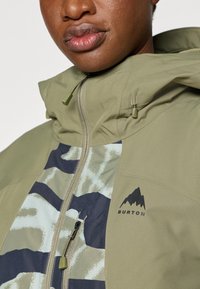 Green waterproof jacket with a hood, featuring a front zip, camouflage pattern in blue and light green, and a black Burton logo on the chest.