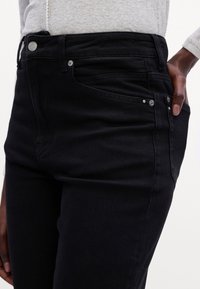 Gros plan d'une personne portant un jean noir près du corps et un haut à manches longues gris clair, la main reposant dans la poche avant.