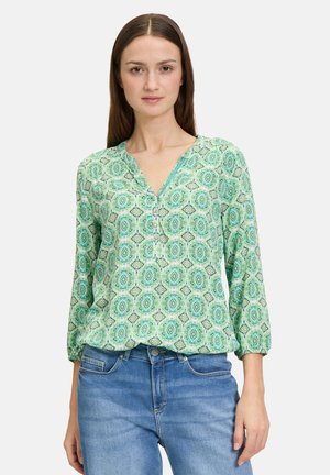 Grüne blusenshirt mit V-Ausschnitt, dreiviertellangen Ärmeln und Knopfdetails, das kreisförmige und geometrische Muster in unterschiedlichen Farbtönen aufweist.