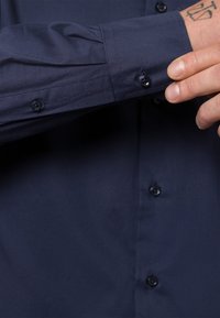 Mörkblå knapp-up skjorta med en slät textur, som har mörka knappar, en knappad manschett och en subtil logotyp på ärmen.