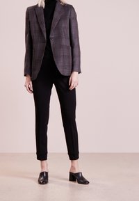 Karierte Blazer in dunklen Tönen, taillierte Passform; kombiniert mit schwarzen, schmalen Hosen und schwarzen, hochhackigen Mules, vor einem hellen Hintergrund.