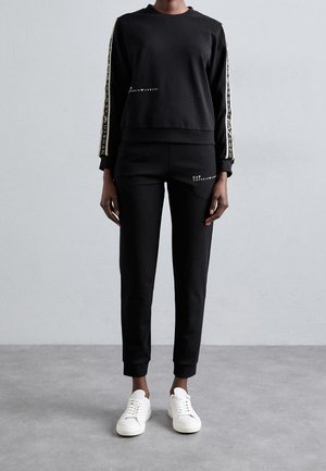 Person mit schwarzem langärmligem Oberteil und Hose mit weißem Logotext auf den Ärmeln und am Oberschenkel der Hose, kombiniert mit weißen Turnschuhen, steht auf grauem Boden.