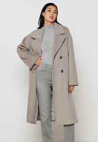Beige wollen mix dubbelzijdig jasje met grote reverskraag, zijzakken en een relaxte pasvorm, gedragen over een grijze coltrui en lichte spijkerbroek.