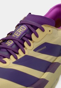 Chaussure de sport violette et beige avec une tige texturée, de larges bandes violettes et des lacets prononcés. Comprend un détail adhésif sur la tige.