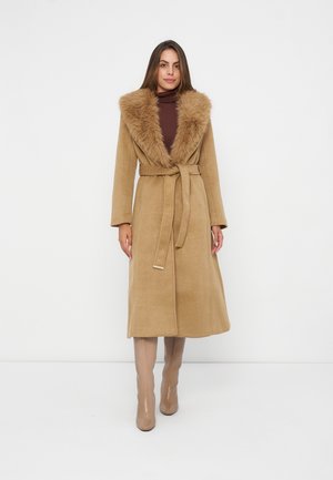 Mujer de pie con un abrigo largo camel con cuello de piel, cintura ceñida, jersey de cuello alto marrón y botas beige hasta la rodilla sobre un fondo blanco liso.