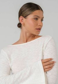 Weißes Oberteil mit langen Ärmeln und weitem Bootsausschnitt, ausgestattet mit lockeren Glockenärmeln. Der Stoff hat eine strukturierte Optik. Goldene Creolen ergänzen das Design.