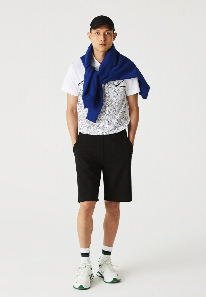 Polo shirt blanc à motifs, shorts noirs, sweat-shirt bleu drapé sur les épaules, casquette noire, baskets blanches avec accents verts, et chaussettes rayées.