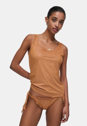 Donna con capelli scuri raccolti che indossa un top bronzo lucido e slip bikini abbinati con lacci laterali su sfondo chiaro uniforme.