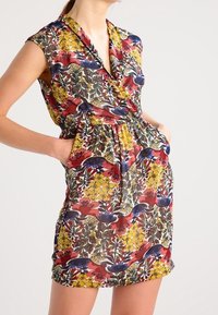 Sukienka w kwiatowy wzór z krótkimi rękawami, głębokim dekoltem w kształcie V i bocznymi kieszeniami; kolory to czerwony, żółty, niebieski i zielony na jasnym tle.