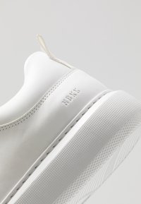 Sneaker bianco in pelle con finitura liscia, caratterizzato da testo in rilievo "NBKK", una suola strutturata e un design minimalista.