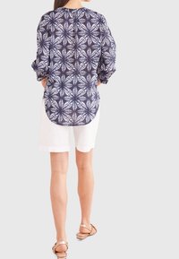Blusa a maniche lunghe blu navy con motivo a foglia, orlo arrotondato e dettagli con bottoni sul retro. Abbinata a pantaloni corti bianchi e sandali.