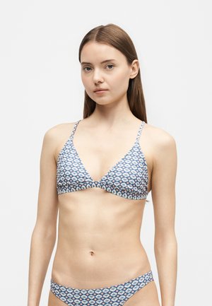 Jeune femme aux longs cheveux bruns portant un haut et un bas de bikini bleu et blanc à motif floral, regardant vers l'avant sur un fond clair uni.