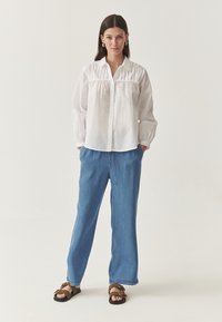 Camicia bianca con dettagli arricciati, abbinata a jeans a gamba larga blu chiaro e sandali marroni. Tessuto morbido, vestibilità rilassata, stile casual.