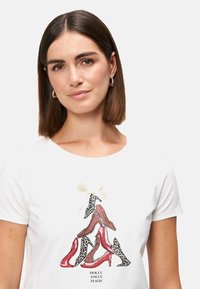 T-shirt blanc arborant un motif de chaussures à talons hauts en rouge, marron et imprimé léopard, orné d'un petit nœud crème au-dessus du design.