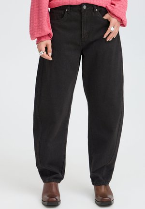 Person iført sorte løstsiddende jeans, brune ankelstøvler, en lyserød tekstureret langærmet top og sølvringe på begge hænder.