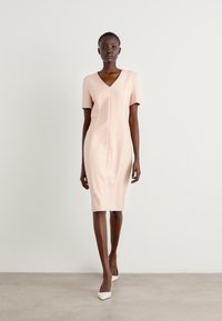 Light pink fitted dress with short sleeves, V-neckline, and a knee-length hem. Smooth texture with a central seam detail. White shoes.