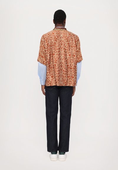 Camisa de manga corta de tela de colores naranja y crema, combinada con una camisa de manga larga azul y pantalones de mezclilla oscuros. Zapatillas blancas completan el look.