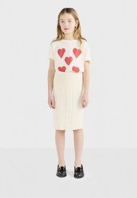 Lys beige skjorte med røde hjerteprint, kombinert med et plissért beige skjørt og svarte sko. Enkelt design, glatt stofftekstur.