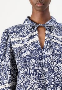Mørkeblå bluse med hvitt blomsterprint, korte puffermer, krage og knyte-lukking. Påfallende blonderdetaljer langs brystet.