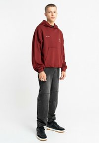 Bordowa bluza z kapturem z przednią kieszenią i logo, połączona z ciemnoszarymi dżinsami jogger z detalami przeszycia oraz czarnymi sneakersami na gumowej podeszwie.